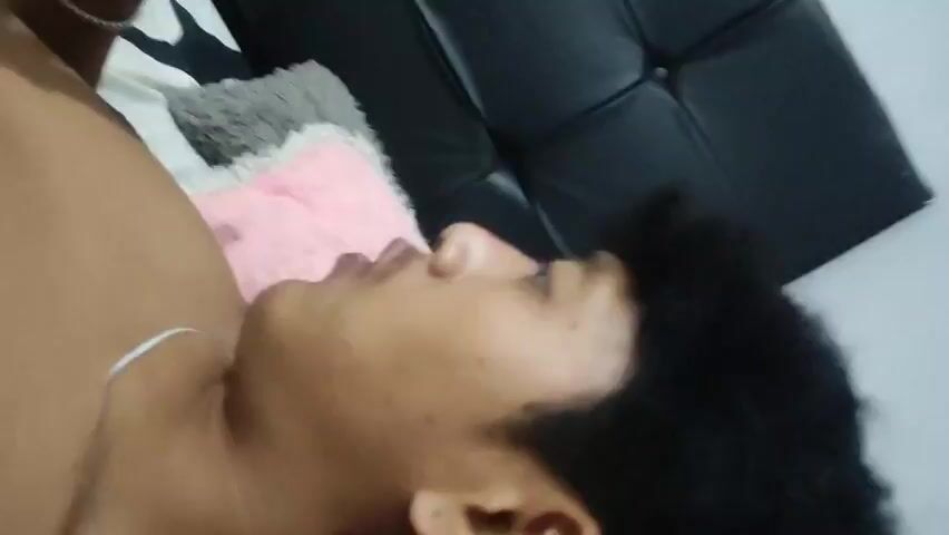 A man lies on his back on a bed with pillows. He is shirtless and wearing a silver chain around his neck. His head is turned to the side, and he appears to be speaking. A black headboard is visible behind him.