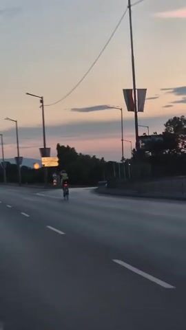 The video captures a cyclist on a road at dusk. The cyclist, wearing a yellow shirt and a helmet, is the primary subject, riding on the right side of the asphalt road. The setting is an urban or suburban area with traffic lights, street lamps, trees, and road signs visible. A Bulgarian flag is prominently displayed on a tall pole. In the background, there are billboards, vehicles, and buildings. The sky displays the soft colors of sunset, with hues of pink, orange, and blue, indicating it's either early morning or late evening. The weather appears clear.

Road signs provide some location context. One sign points towards "Airport Sofia" and also indicates a speed limit of 70. Another sign has Cyrillic text which is partially obscured but appears to be an advertisement. The overall mood is peaceful and transitional, reflecting the quiet moments of dusk.