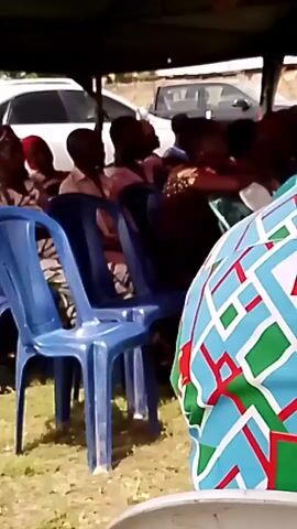 A medium shot focuses on a group of people seated in plastic chairs under a green canopy. The chairs are blue, and some are empty. The people appear to be gathered for an event, possibly a political rally given the "APC" logo visible on the back of a person's shirt in the foreground. Several individuals are partially visible, wearing patterned clothing and headwraps. In the background, white cars are parked under the canopy. The ground is covered in grass.