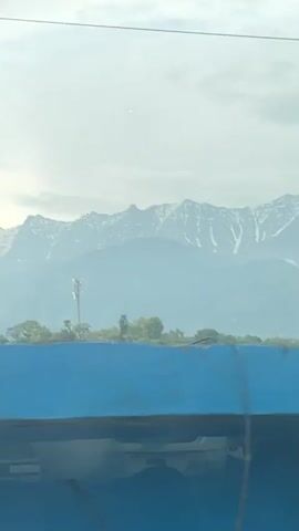The video shows a mountain range with snow-capped peaks under a cloudy sky. The camera is likely inside a moving vehicle, and the foreground contains various objects, including a building roof, a billboard with the text "30 YEARS OF TRUST", and a tree. The camera moves, causing the foreground objects to shift, providing different perspectives of the mountain range in the background.