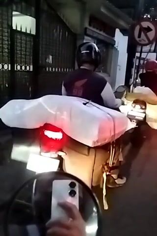 A first-person perspective video shows a motorcycle rider in traffic. The rider is wearing a helmet and a dark jacket with a red logo. They are carrying a large white bag on the back of their motorcycle, along with other items. Traffic is congested, with several other motorcycles and a silver car visible. The car has a license plate that reads "S 1417 FZ". A traffic sign indicating "no right turn" is visible on the side of the road. The video appears to be shot during the daytime, with trees and buildings lining the street.