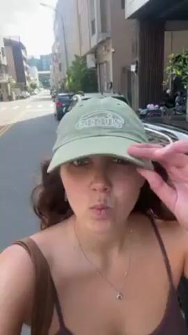 A young woman with light skin, curly brown hair, and a light green baseball cap is taking a selfie video on a city street. She is wearing a brown tank top and a necklace. She makes a variety of faces at the camera, including pouting, sticking her tongue out, smiling, and puckering her lips. Behind her, there are parked cars and scooters lining the street. Buildings are visible in the background. The video appears to be taken during daylight hours.