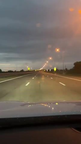 The video shows a car driving down a wet highway at dusk.  The sky is overcast. The car's headlights illuminate the road, and streetlights line both sides of the highway. Road markings are visible on the pavement.  There is sparse traffic ahead.  The view is from the driver's perspective, looking out through the windshield.