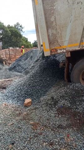 A dump truck unloads a pile of small, gray stones. The truck bed is angled upward, and the stones are pouring out of the back onto a growing heap. The truck is dirty and has yellow reflective strips. The ground around the pile is covered in similar stones and some dirt and sticks. In the background, a construction worker wearing a red hard hat and yellow safety vest stands near a wooden fence and other construction materials. Another person is visible further back, standing near some trees. The sky is overcast.
