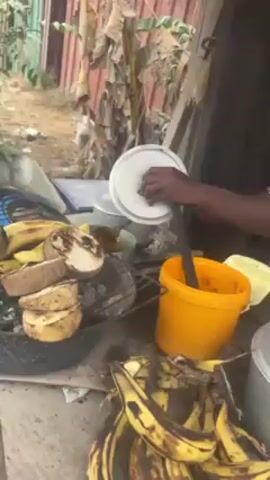 The video shows a woman in a floral shirt and white head covering standing at a makeshift outdoor kitchen preparing food. She is using a lid to scoop something from a pot into an orange container. There are ripe plantains on the table. The camera then moves to a man in a white "Givenchy" shirt standing outside. The scene shifts to show a cluttered, dirt-filled area with shacks and temporary structures, suggesting a poor, possibly rural environment. In the final shot, a road construction site is visible in the background, with piles of wood and construction materials scattered nearby. A person wearing a high-visibility vest is working on the road.