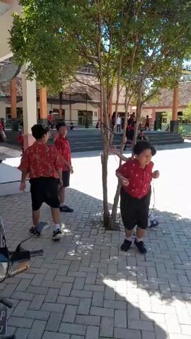 A group of young boys in red shirts and black shorts are gathered outdoors in a paved area. Some are standing, while others are walking or running. A tree with green leaves is in the center of the frame, and the boys are interacting around it. In the background, there's a building with an orange roof and a covered walkway. A motorcycle is partially visible in the lower left corner. The overall scene appears to be a schoolyard during the daytime.