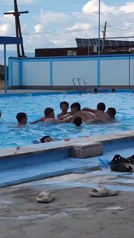 A group of young males are interacting in a swimming pool. Some are in the water, while others are partly submerged. A few individuals are clustered together, appearing to be in a playful or wrestling interaction. The pool has blue tile edging, and the surrounding area is concrete. Several pairs of flip-flops are scattered on the concrete. In the background, a white and blue building with a ladder extends into the pool. The sky is visible with clouds.