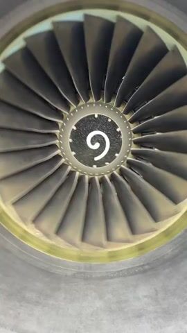 The video shows the front of a static jet engine. The blades are visible and spinning at a low rate of speed, revealing a black circle at the center with a white spiral design. The engine casing is gray and appears to be constructed of riveted metal panels. There are no people or other objects visible in the video. The setting appears to be an aircraft maintenance facility or airfield.