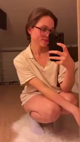 A young woman in her twenties, with light brown hair pulled back and round glasses, is taking a selfie in front of a mirror. She's squatting on a white, fluffy rug on a light wood floor. She's wearing a light beige short-sleeved shirt and what appear to be shorts or underwear of the same color. She's smiling slightly at her reflection while holding a black smartphone. The room has a sloped ceiling, suggesting it might be an attic. A metal railing is visible behind her, and a wooden piece of furniture is partially seen in the background to the right. The lighting is soft and warm, possibly from artificial sources.