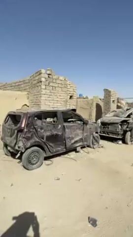 The video shows a desert-like environment with damaged buildings. In the foreground, there are two destroyed cars. Three men on a motorcycle drive past the damaged vehicles, further into the town. One man in front carries a rifle. The sky is clear and blue.