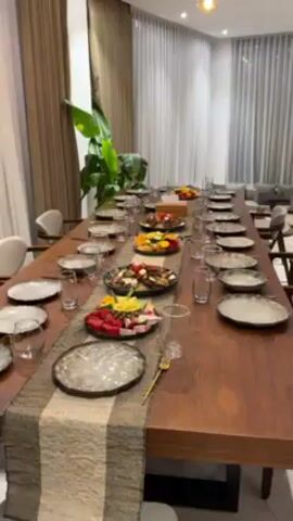 The video shows a long, wooden table set for a meal, with place settings for numerous people. Each setting includes a plate, a glass, and a gold-colored fork. A long, neutral-toned table runner stretches down the center of the table. At intervals along the runner, there are platters of food; some platters contain fresh fruit, while others appear to hold a meat dish with rice and vegetables. A large, leafy plant stands near the end of the table. The room has sheer curtains covering the windows, and there are upholstered chairs at the table. The lighting appears to be warm and inviting. No people are visible in the shot.