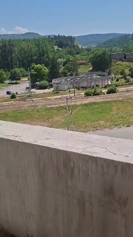 The video pans across a landscape featuring rolling green hills, trees, and a clear blue sky. In the foreground, a rough concrete wall is visible. Below the wall, train tracks run horizontally across the frame. Further in the distance, a small building with a distinctive wavy roofline sits next to the tracks. Beyond that, more trees and buildings can be seen, with hills forming the background. To the right, a paved road runs parallel to the tracks, with parked cars visible on the side. A Bulgarian flag is also seen flying from a flagpole near the road. The overall impression is of a rural or semi-rural setting on a sunny day.