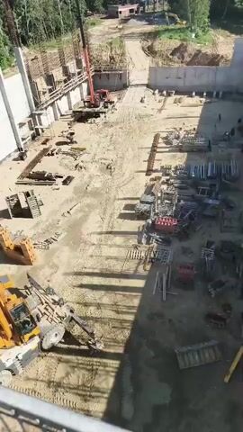 This aerial video captures a construction site on a sunny day in Primorye, Russia. The scene is dominated by unfinished concrete structures, including tall pillars and walls, interspersed with scaffolding. The ground is a mix of dirt and sand, with tire tracks indicating the movement of heavy machinery.

Numerous construction materials are scattered across the site, including stacks of metal bars, wooden pallets, and various metal frames. A large orange crane is positioned near the center of the frame, suggesting ongoing work. In the foreground, a yellow excavator is parked, its mechanical arm visible. A few individuals can be seen working on the site, though their activities are not clearly discernible from this distance.

The background reveals a natural landscape with a dense forest of green trees under a clear blue sky with scattered white clouds. In the distance, some smaller structures and a paved road can be observed, hinting at the surrounding environment. The overall impression is one of active construction and development within a semi-rural setting. There is no visible text in the video.