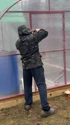 A man in a camouflage jacket and dark sweatpants is using a cordless drill to attach a transparent corrugated panel to a greenhouse. He tightens a screw, takes a step back, and adjusts the hood of his jacket while looking directly at the camera. The greenhouse has a red frame, and the base is made of wooden planks. The ground around the greenhouse is covered in grass and a few boxes are sitting on the ground. The overall setting appears to be a backyard or garden.