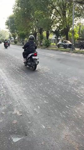 The video shows a street scene with several motorbikes. In the foreground, a person on a dark-colored motorbike is riding away from the camera. They are wearing a dark jacket, helmet, and pants. The motorbike has a license plate and a red taillight. The road is paved and appears to have some debris or rough patches. In the background, several other motorbikes are visible, along with trees and some parked vehicles. The scene appears to be in daylight.