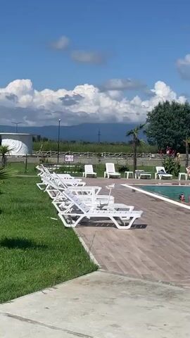 The video shows an outdoor swimming pool area on a sunny day with scattered clouds. In the foreground, several white lounge chairs are positioned on a wooden deck. The pool itself is filled with clear turquoise water, and several people are swimming and interacting within it. Some individuals are partially submerged, while others are more visible. On the deck near the pool's edge, there are a few red and orange objects, possibly pool toys or flotation devices. Beyond the pool area, a grassy lawn is visible, also lined with white lounge chairs. Further in the background, a wooden fence separates the pool area from a field. A line of trees forms a backdrop, with mountains visible in the distance. A single palm tree stands near the pool. Two individuals are visible relaxing on lounge chairs in the background, and another person is standing by the pool. Several other people are seen walking or standing around the pool area. The overall scene depicts a recreational setting, likely a resort or a public pool, with people enjoying leisure activities.