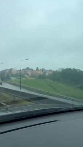The video shows a view from inside a vehicle, looking out through a wet windshield. It appears to be raining. The windshield has water streaks and raindrops on it, obscuring the view somewhat. In the distance, there are buildings and trees. A road or highway is visible, with a guardrail and a lamppost. A dark-colored car is seen on the road in the distance, moving away from the camera. The sky is overcast and gray. The overall impression is of a gloomy, rainy day.
