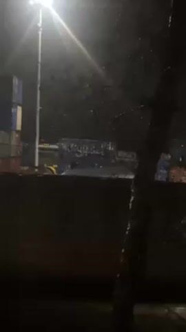 The video shows a dimly lit outdoor scene at night, possibly an industrial or port area. The camera is stationary, looking out from what appears to be an indoor location with some water droplets on the surface, possibly a window. In the distance, there are stacks of shipping containers, some marked with the word "JIEAN". A bright street light illuminates the scene, casting a stark glow and creating long shadows. A forklift is also visible, although details are obscured by the distance and low light. A bare tree is visible in the foreground on the right side of the frame, partially obscuring the view. The overall impression is one of a quiet, slightly eerie, and industrial nighttime environment.
