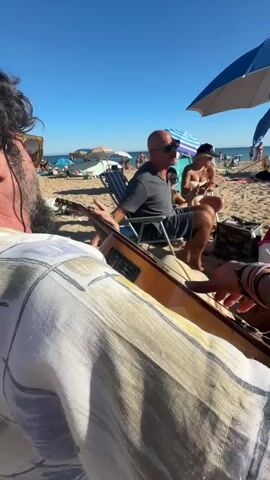 The video shows a close-up of a person playing an acoustic guitar on a beach. In the background, a bald man in a grey t-shirt and black shorts is sitting in a beach chair, wearing sunglasses, and clapping his hands. To his right, a woman with blonde hair is also sitting in a beach chair, wearing a white bikini top and sunglasses, and appears to be listening. The beach is populated with other beachgoers, umbrellas, and beach chairs. The sky is clear and blue, indicating a sunny day.