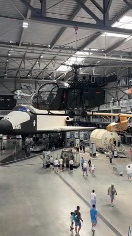 The video shows a wide interior view of a large, industrial-looking building, likely a museum or exhibition hall. The ceiling is high and industrial, with exposed metal beams and lighting fixtures. The floor is a smooth, gray surface, likely concrete.

In the center of the frame, a space shuttle is prominently displayed, suspended from the ceiling. It is white with black markings. Attached to the underside of the space shuttle is a dark-colored helicopter, also suspended. The helicopter has German military markings.

To the right of the space shuttle, a cylindrical structure, possibly a space module or habitat, is visible.

Numerous people are scattered throughout the space, walking around and observing the exhibits. They are dressed in casual summer clothing. Some appear to be families with children.

Further to the left and in the background, other exhibits are visible. These include various aircraft, including what appears to be a biplane hanging lower than the other exhibits, and a shiny, dark-colored jet with its nose pointing downwards. Several vintage vehicles, including red fire trucks, a silver caravan, and green utility vehicles, are also on display.

The overall impression is of a vast exhibition space showcasing a diverse collection of historical aircraft, spacecraft, and vehicles. The lighting is bright and even, illuminating the exhibits and the visitors.