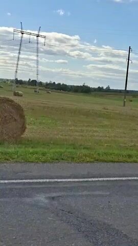 The video captures a rural landscape with a field under a partly cloudy blue sky. In the foreground, a portion of an asphalt road with a white line is visible. Beyond the road, a grassy field stretches out, with several round hay bales scattered across it. Two tall power line towers stand in the field, with wires running between them. One of the power line poles is leaning significantly. The overall impression is of an agricultural area on a clear day, with signs of farming activity.