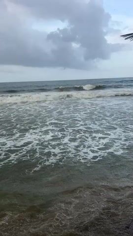 The video shows a cloudy sky over a beach. Waves are crashing on the shore. The water is a murky green and brown. The sand is a light brown. There are no people or animals visible in the video.