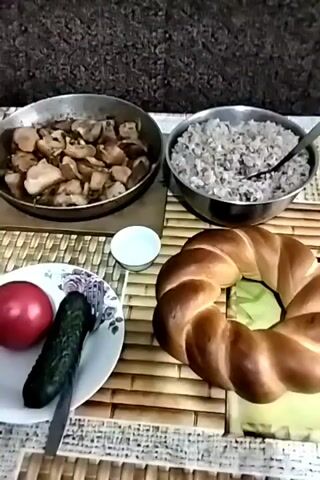 The video shows a meal laid out on a bamboo placemat. In the upper left is a round, shallow pan filled with cooked meat, appearing to be stewed or braised. Next to it, to the right, is a round metal bowl filled with a grainy, cooked dish, possibly rice or buckwheat. Below the pan of meat and to the left is a white plate with a red tomato and a green cucumber, along with a fork. In the center, between the plate and the bowl of grain, is a small white dish. To the right of the grain bowl is a large, circular loaf of braided bread. The background is dark and out of focus, suggesting an indoor setting. The video consists of a single static shot with no discernible action or interaction.