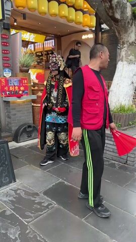 Here is a detailed description of the video:

The video shows a person in a traditional Chinese costume standing outside a shop. The costume includes a red cape, a black robe embroidered with golden dragons, matching pants, and an elaborate headpiece. The person also wears a mask covering the upper part of their face. The person is holding two red fans and poses for the camera, making a V-sign with their fingers. 

The background features a shop with yellow lanterns hanging outside, and a few people are walking by. The surrounding area appears to be a paved street with some greenery and decorative elements.