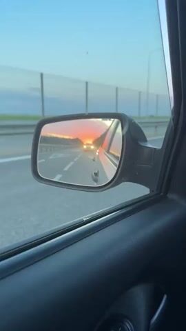A camera records a passenger side mirror showing a sunset. The sun is a bright orange color in the distance on a two-lane road with a dotted white line between the lanes. A car approaches with headlights on. The car's driver side is a silver color. Another vehicle drives by on the left side of the road. A metal fence is on the left side of the road. Trees are in the distance. The vehicle's interior is black.