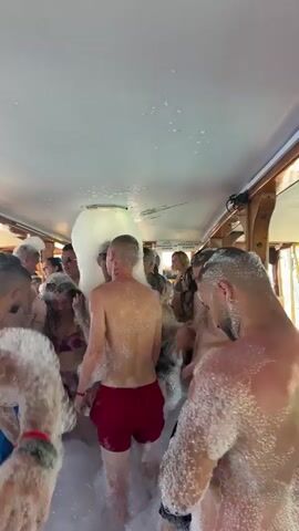A group of people, both male and female, are enjoying a foam party inside what appears to be a boat or a covered venue. Most individuals are shirtless or wearing swimwear, such as bikinis and swim trunks, and are covered in white foam. A foam machine is visible at the back, actively dispensing more foam into the crowded space. The interior features a white ceiling and wooden accents on the walls, with light from outside filtering through windows along the sides, indicating it is daytime. Participants are smiling and laughing, engaging with the foam and each other, suggesting a lively and fun atmosphere. The event is taking place in Menderes, Turkey.