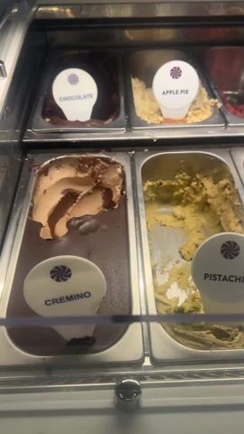 The video shows a refrigerated display case containing multiple tubs of gelato. The tubs are labeled with flavors such as chocolate, apple pie, strawberry, mango sorbet, Dubai, lemon, cremino, pistachio, yogurt with berries, blackcurrant cheesecake, and Ferrero Rocher. The gelato appears to be at different levels of consumption, indicating varying popularity of the flavors. The display case is made of stainless steel with a transparent front panel.