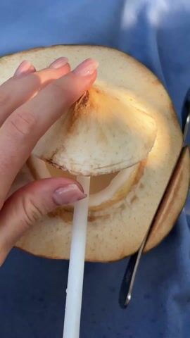 Here's a detailed description of the video:

The video shows a close-up shot of a partially opened young coconut on a blue surface. A hand with polished fingernails lifts the cut-off top portion of the coconut, revealing the white flesh inside. A white straw is inserted into the coconut's liquid. A knife is seen to the right of the coconut on the blue surface. The coconut's exterior is a light yellowish-green. Some fibers are visible around the opening. The inside of the coconut is filled with a translucent liquid. The lighting suggests an outdoor setting.