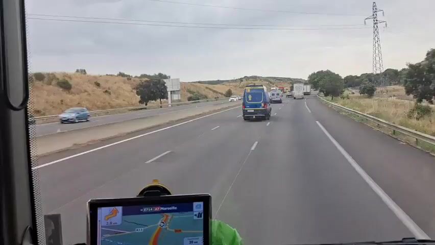 The video shows a view from inside a vehicle, likely a truck or van, as it travels on a highway. In the foreground, a GPS device is mounted on the dashboard, displaying a map with a route to Marseille. The vehicle is in the right lane, with other vehicles ahead, including cars and trucks. A blue van with yellow markings and the text "GENDARMERIE" is visible in the middle lane, passing by. The landscape outside the vehicle is dry and hilly, with some scattered trees. The sky is overcast. The video captures movement along the highway, indicating travel.