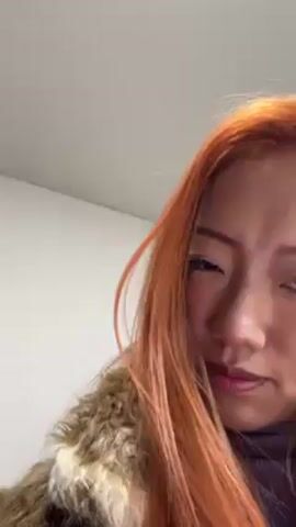 A close-up video features a woman with long, orange hair. She is wearing a purple turtleneck sweater and a furry jacket. The woman displays her long, white, pointed nails, curling her fingers into a claw-like shape. The background consists of a white wall and a glimpse of a white ceiling.