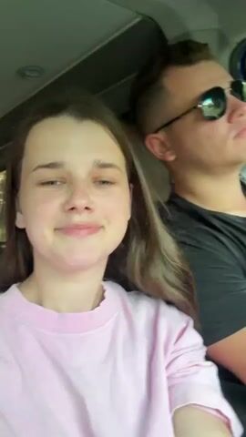 This is a close-up selfie video of a young couple in a car. The woman is in the foreground on the left, wearing a pink sweatshirt. She has brown hair and is smiling at the camera. She has freckles on her cheeks and nose. To her right and slightly behind her is a man wearing a black t-shirt and sunglasses. He is looking upwards and to the right, with his mouth slightly open. He has short brown hair. They are both wearing seatbelts. The scene appears to be daytime, with greenery visible through the car window, suggesting they are driving through a rural or suburban area. The mood seems relaxed and happy. There is no visible text in the video. The timestamp "00:00" is present in the bottom left corner of the video frame.