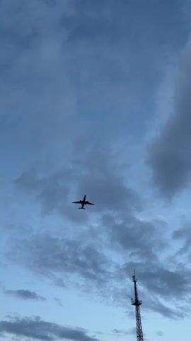 A silhouette of a passenger airplane is seen flying in the sky against a cloudy blue sky. The plane is in the center of the frame, appearing to be in descent. The underside of the plane is visible, with its wings extended and engines under each wing. The plane has landing gear extended. A tall, thin structure resembling a radio tower or antenna is visible in the lower right corner of the frame. The clouds are scattered and appear to be a mix of white and gray. The overall lighting suggests it may be dawn or dusk.