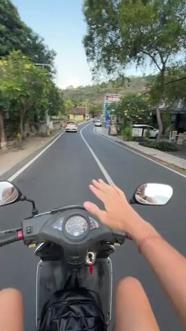 A first-person perspective video shows a person riding a scooter down a paved road lined with trees and buildings. The rider's hand is resting on the scooter's handlebars, and a bracelet is visible on their wrist. Several cars and other scooters are on the road ahead. The sky is clear and blue, with some clouds visible. The scenery suggests a tropical or subtropical location.
