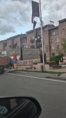 The video shows a drive through a town with buildings and the Armenian flag displayed. The first vehicle observed is an old orange water truck. Then, various other vehicles pass by, including a silver minivan, a green station wagon, and a silver sedan. Buildings line the street, and the sky is cloudy. There is a green bug sculpture in the median. The perspective is from inside another vehicle, showing the side mirror.