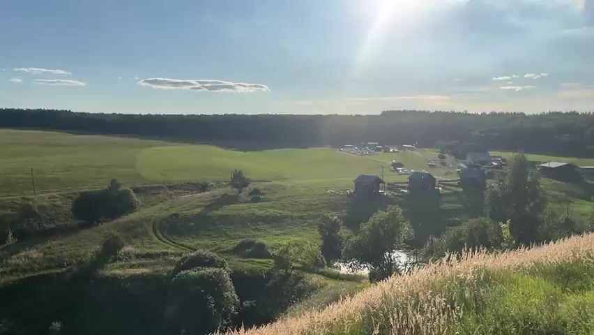 The video captures a rural landscape under a bright sky with sunlight streaming in from the right. The foreground features a gentle slope covered in green grass with scattered trees. In the mid-ground, several small wooden structures, possibly cabins or sheds, are situated on the elevated terrain. Some appear to have porches. Beyond these structures, a larger open green field stretches out. In the distance, a dense forest forms a dark green backdrop. A cluster of buildings, including what appear to be white rectangular structures and darker barns, is visible within the field, near the edge of the forest. There are also some fences and what might be vehicles or equipment present at the distant buildings. Smoke or steam is visible rising from one of the mid-ground structures, and possibly from the distant buildings as well. The overall impression is of a quiet, pastoral setting.