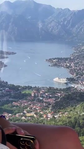 A vertical video shows an aerial view of a large body of water, possibly a bay or fjord, surrounded by a city and mountains. A large white cruise ship is docked in the water near the city. Several small boats leave white wakes on the water. The mountains are a hazy blue. In the foreground, the legs of a person holding a black and gold smartphone are visible, suggesting the video was filmed from a high vantage point, possibly a cable car or a lookout.