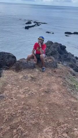 A medium shot shows a person sitting on a rocky outcropping, overlooking the ocean. The person wears a red t-shirt, patterned shorts, gray Crocs, a fanny pack, and a blue and yellow helmet. They have long blonde hair and glasses. The ground is reddish-brown and rocky. In the distance, the ocean stretches to the horizon with scattered dark rocks visible near the shoreline. The sky is overcast. The camera slowly zooms in on the person.