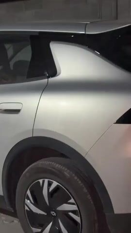 A silver car is parked in a driveway at night. The car is a four-door sedan with tinted windows. The front of the car is not visible, but the side of the car is visible, including the front door, the rear door, and the rear wheel. The car is parked in front of a brick wall. The car appears to be in good condition. There are no people visible in the video.
