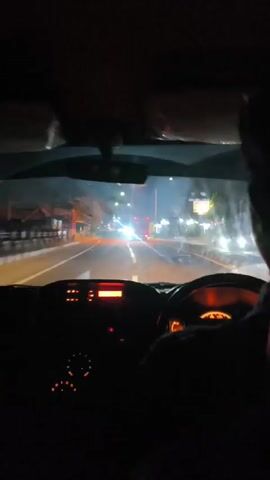 The video shows a first-person view from inside a car driving at night. The dashboard lights are illuminated, and the steering wheel is visible. Ahead, the road is lit by streetlights and the headlights of other vehicles. A red truck is seen in the distance, driving in the same direction. The scenery outside includes buildings and trees lining the road. The camera perspective suggests the viewpoint of a passenger or driver looking forward through the windshield.