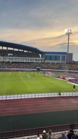 A soccer game is taking place on a green field under a cloudy sky. Spectators are seated in the stands, and some are standing near the field. The sun is setting, casting a warm glow on the scene. A scoreboard is visible on the right side of the field, and a large light tower is also present. There are players in white and green uniforms on the field, and a goalkeeper is in position in front of the net. The atmosphere appears to be one of excitement and engagement.