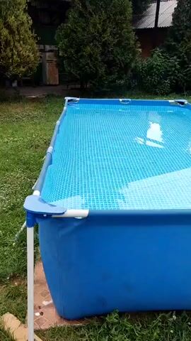 A medium shot captures a rectangular, above-ground swimming pool filled with clear blue water. The pool's frame is white, and its sides are a vibrant blue fabric. The water's surface has a slight ripple, reflecting the sky and surrounding greenery. A white support leg is visible on the left side of the pool.

The pool is situated outdoors on a grassy lawn. In the background, lush green trees and bushes create a natural backdrop. A hint of a building with a dark roof can be seen behind the foliage, suggesting a residential area or a yard. The overall scene appears to be a peaceful, sunny day, likely in the late morning or afternoon, given the brightness and the shadows. The setting evokes a sense of summer leisure and relaxation, common in suburban or rural environments during warmer months. No people are visible in the frame, emphasizing the tranquil solitude of the pool. There is no visible text in the media file.