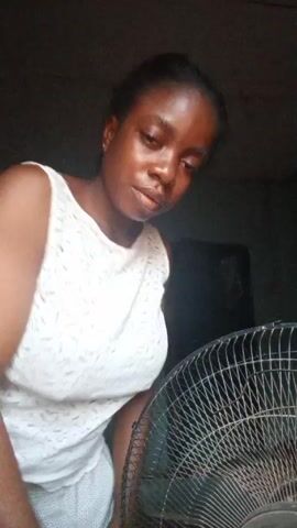 A woman stands indoors in front of an electric fan. She wears a white sleeveless lace top. She is facing forward, looking down slightly at the camera. She puts her hands on her face briefly. The background is dark.