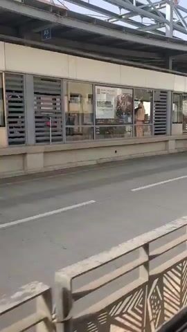 The video shows a view from a moving vehicle looking towards a bus station or transportation hub. The station has a modern design with a white facade, large windows, and a metal roof structure. Inside the station, there are visible signs, advertisements, and what appear to be ticket booths or waiting areas. Some windows show reflections, obscuring the view of people inside. There are turnstiles visible in some sections. A road with lane markings is in the foreground, and a metal railing separates the road from the viewing perspective. The overall environment appears to be an urban or suburban setting.