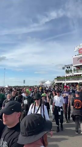 The video shows a large crowd of people walking around an outdoor event on a sunny day. A large stage is visible in the background, with a performance taking place. There are also several large screens displaying the performance, and various brand banners and structures, including a Jim Beam observation deck. People are dressed in casual attire, some wearing band t-shirts or hats. Some individuals are holding drinks. The ground is paved, and the sky is blue with some clouds.