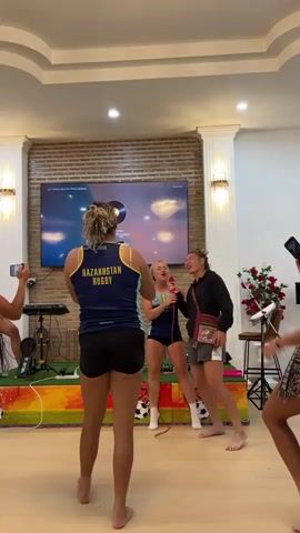 The video shows a group of young women inside a room, possibly a common area or recreational space. In the foreground, a woman with blonde hair in a ponytail faces away from the camera, wearing a dark blue sleeveless top with "KAZAKHSTAN RUGBY" printed on the back in yellow and white lettering, and black shorts. She appears to be observing or participating in an activity. To her right, two other women are more actively engaged. One woman in a dark jacket and grey shorts is raising her arm in a celebratory gesture while singing into a microphone. She has a patterned bag slung across her body. Next to her, a woman in a dark blue outfit and dark shorts is also singing into a microphone and laughing. She is wearing white socks. Behind them, a large television screen displays what appears to be a music video or karaoke interface with some sort of abstract imagery. The room has a modern aesthetic with a brick wall behind the screen and decorative pillars. There are also plants and what looks like musical equipment in the background. Other individuals are partially visible, with one person on the far right also wearing a similar dark blue and yellow top, and another person on the far left holding up a phone, likely recording. The floor is a light-colored wood.