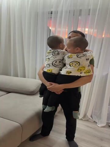 A man is holding two young children, who appear to be twins. The children are wearing matching t-shirts with smiley face designs. The man is standing in a room with a large couch and sheer curtains. The lighting suggests it is either dawn or dusk. In the background, another child is lying on the couch.